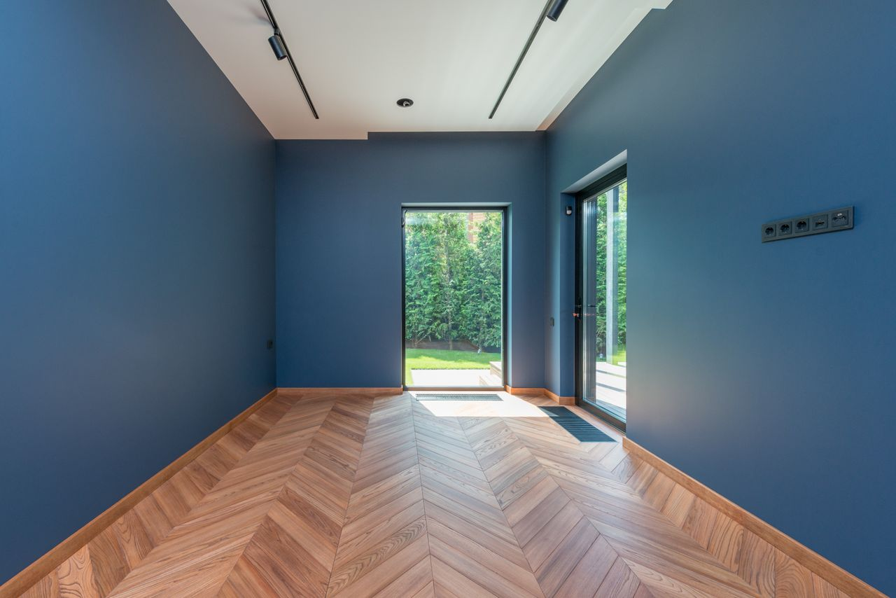 about-img Spacious room featuring blue walls, wooden herringbone floor, and glass doors leading to a garden.